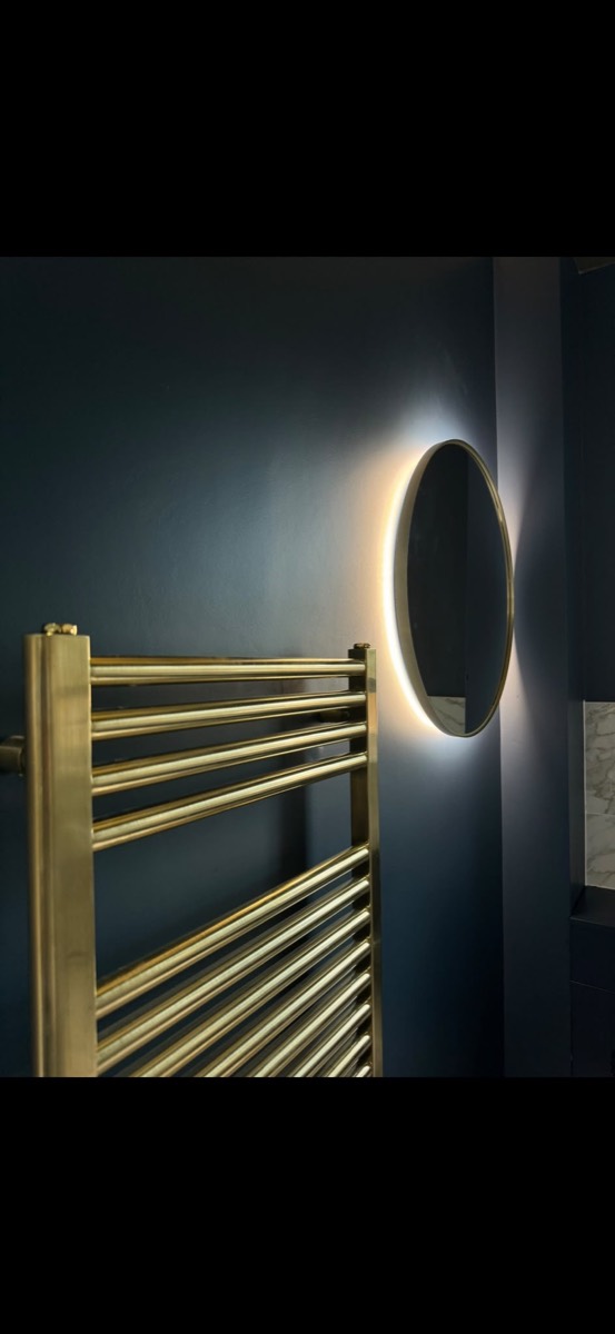 Brushed brass heated towel rail and a backlit round mirror on a deep navy bathroom wall